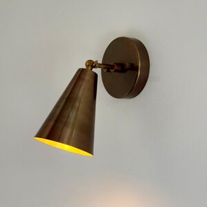 May include: A bronze-coloured wall sconce featuring a conical shade and a circular backplate. The interior of the shade is yellow. The sconce is designed to be adjustable. The fixture is made of metal.