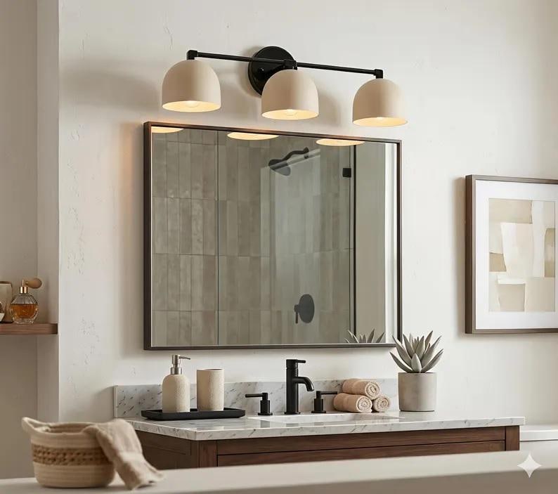 Puede incluir: Un tocador de ba&ntilde;o moderno con un espejo rectangular grande y una l&aacute;mpara de tres luces. La l&aacute;mpara tiene un marco negro y pantallas beige en forma de c&uacute;pula. El tocador cuenta con una encimera de m&aacute;rmol, un mueble de madera y accesorios negros.