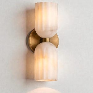 May include: A wall-mounted light fixture with two alabaster-colored, cylindrical shades. The shades have vertical grooves and are connected by a brass-colored circular base. The fixture is designed for indoor use and provides soft, ambient lighting.