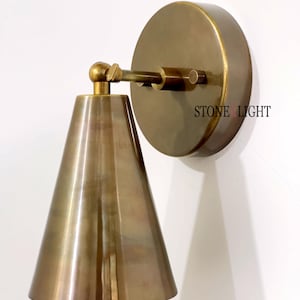 May include: A brass wall sconce featuring a conical lampshade and a circular backplate. The sconce has a pivoting arm and is mounted on a white wall. The text "STONE LIGHT" is visible on the backplate.
