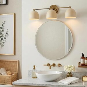 Puede incluir: Un interior de ba&ntilde;o con un espejo redondo, una l&aacute;mpara de tocador de tres luces y un lavabo blanco. Las luces del tocador tienen pantallas beige y un acabado de lat&oacute;n. Tambi&eacute;n se ven una impresi&oacute;n bot&aacute;nica enmarcada y una caja de almacenamiento tejida.