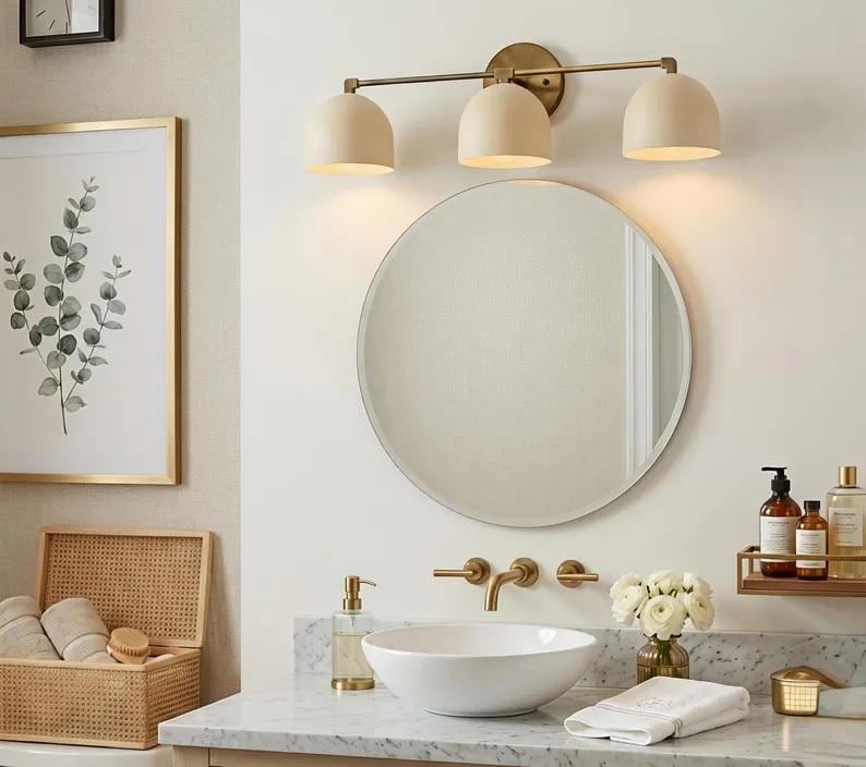 Puede incluir: Un interior de ba&ntilde;o con un espejo redondo, una l&aacute;mpara de tocador de tres luces y un lavabo blanco. Las luces del tocador tienen pantallas beige y un acabado de lat&oacute;n. Tambi&eacute;n se ven una impresi&oacute;n bot&aacute;nica enmarcada y una caja de almacenamiento tejida.