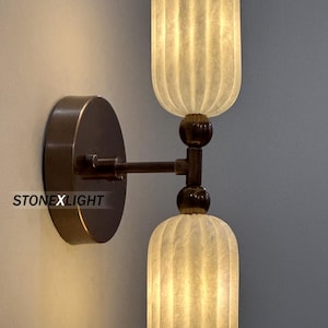 Può includere: Applique da parete con due paralumi in stile alabastro. I paralumi hanno un design a coste ed emettono un bagliore caldo. L'apparecchio ha una finitura bronzo. Il testo "STONEXLIGHT" è visibile.