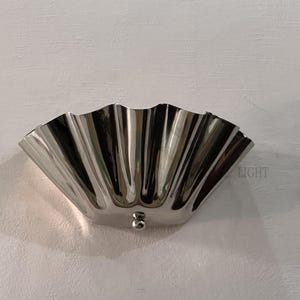 May include: A polished, silver-toned wall sconce with a scalloped, fan-like design. The fixture has a small, round detail at the bottom. The word "LIGHT" is visible on the right side. This decorative lighting fixture is mounted on a white wall.