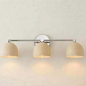 Puede incluir: Una l&aacute;mpara de tocador de tres luces con acabado cromado pulido. Cuenta con tres pantallas beige en forma de c&uacute;pula. La l&aacute;mpara est&aacute; montada en una pared, adecuada para ba&ntilde;os o tocadores.