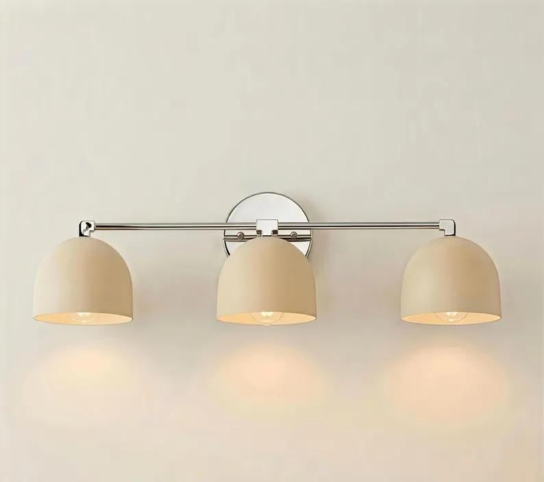 Puede incluir: Una l&aacute;mpara de tocador de tres luces con acabado cromado pulido. Cuenta con tres pantallas beige en forma de c&uacute;pula. La l&aacute;mpara est&aacute; montada en una pared, adecuada para ba&ntilde;os o tocadores.