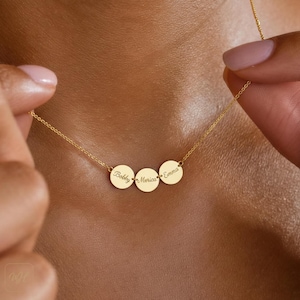 Pode incluir: Um colar dourado com três pingentes circulares. Cada pingente é gravado com um nome: "Bobby", "Merica" e "Emma". O colar é exibido no pescoço de uma pessoa.