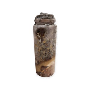 May include: A cylindrical glass bottle with a textured, frosted exterior surface. The bottle features a dark, mottled brown and tan interior content with visible granular particles and layered sediment throughout the body and a dark cap.