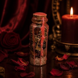 May include: A clear glass bottle sealed with a cork and dripping pink wax, filled with layers of white salt, dark stones, and a gold-colored skeleton key. The bottle is positioned upright, showcasing the internal contents and the wax seal.