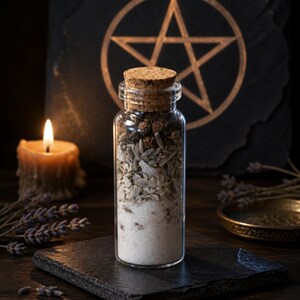 May include: A small glass bottle with a cork stopper, containing layers of white, beige, and brown ingredients. The bottle is placed on a black slate, with a lit candle and a pentagram in the background. The image suggests a spiritual or ritualistic theme.