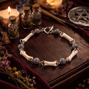May include: A silver chain bracelet featuring bone-shaped beads and gray gemstone accents. The bracelet is displayed on a dark wooden surface, with candles and small glass bottles in the background. The overall aesthetic is gothic and mystical.