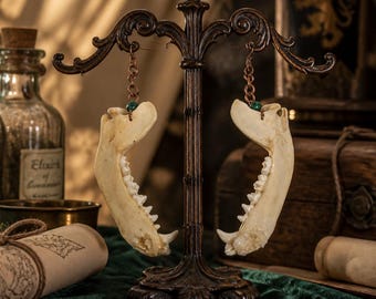 Puede incluir: Un par de pendientes con mandíbulas de animal en color hueso suspendidas de cadenas de metal cobrizo. Cada pendiente incluye una pequeña cuenta redonda de color verde oscuro en la unión, colgados de un soporte de metal oscuro.