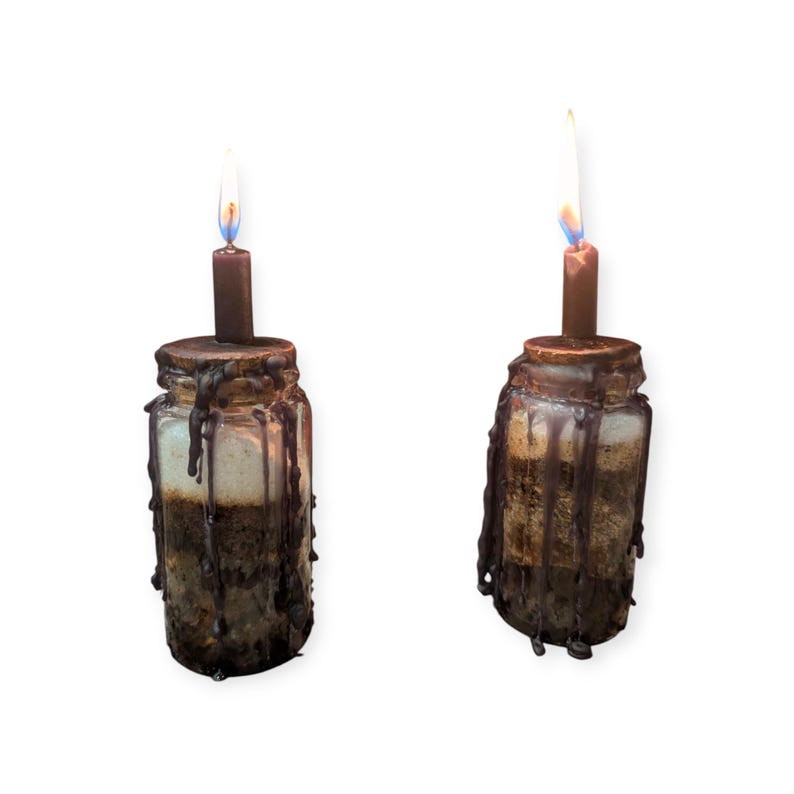May include: Two glass jars filled with layered sand in shades of beige and brown, topped with dark brown wax that drips down the sides. Each jar features a single lit brown candle centred on the lid, creating a warm glow from the flame.