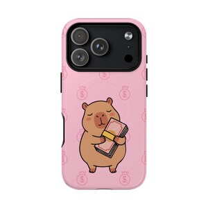 May include: Pink phone case featuring a cartoon capybara holding a stack of money. The capybara is brown with a light brown belly. The background has a repeating pattern of dollar signs and crowns.