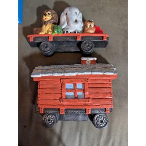 May include: Two painted ceramic train cars. The top car is a black flatbed carrying a brown lion, a green turtle, a gray elephant, and a brown walrus. The bottom car is a red caboose with a white-topped roof, a central window, and black wheels.