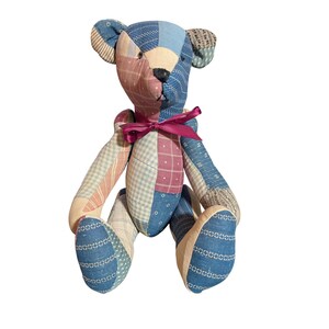 May include: A handmade patchwork teddy bear with a pink ribbon around its neck. The bear is made of various fabrics in shades of blue, pink, and cream. The bear's nose is black, and the ears are a patterned fabric.