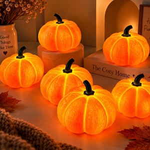 Peut inclure: Un ensemble de six lumières orange en forme de citrouille avec des tiges sombres. Les citrouilles sont disposées sur une surface avec un vase blanc portant l'inscription Good Things Take Time et un livre intitulé The Cozy Home avec un cœur.