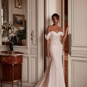 Puede incluir: Un vestido de novia blanco hasta el suelo con escote de hombros caídos, mangas drapeadas, un corpiño ajustado y una falda cubierta de intrincados adornos de lentejuelas y flores que se extiende en una larga cola fluida.