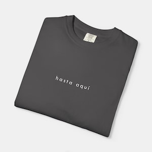 Puede incluir: Una camiseta gris oscuro con la frase "hasta aquí" impresa en blanco. La camiseta está doblada cuidadosamente, con la etiqueta de la marca visible en el cuello. La camiseta está hecha de un material suave y cómodo.