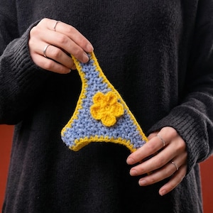 Puede incluir: Un top de bikini de ganchillo azul y amarillo con una flor amarilla en el centro. El artículo se sostiene contra un suéter gris oscuro. El artículo hecho a mano es probablemente para el verano.