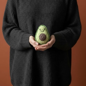 Può includere: Un giocattolo di avocado all'uncinetto con una faccia sorridente. L'avocado è verde con un nocciolo marrone e viene tenuto tra le mani. Lo sfondo è di colore marrone pieno.