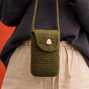 May include: Olive green crochet crossbody phone bag with a flap closure and a white flower accent. The bag has a long strap and is handmade. A stylish and practical accessory for carrying a phone or small items.