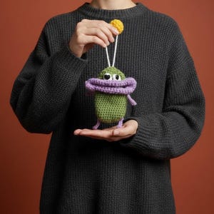 Pode incluir: Uma criatura verde feita à mão em crochê com uma boca roxa e pequenos braços e pernas. A criatura tem olhos brancos e é suspensa por um fio branco com uma bola amarela no topo. O fundo é de cor ferrugem sólida.