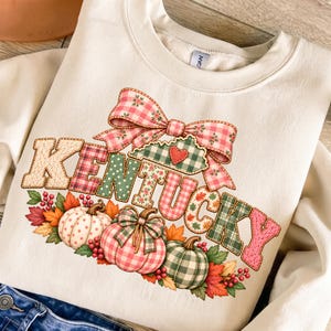 Op de afbeelding: Een crèmekleurige sweater met ronde hals en een grafische print van het woord KENTUCKY in diverse ruit- en stippenpatronen. Boven de tekst staat een grote roze en wit geblokte strik en daaronder drie pompoenen met herfstbladeren.