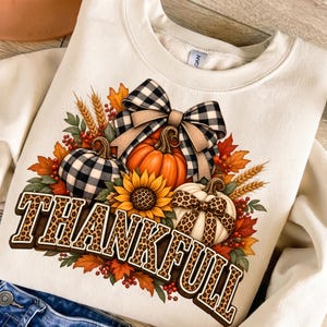 Op de afbeelding: Een crèmekleurig sweatshirt met ronde hals en een afbeelding van drie pompoenen, een zonnebloem en een grote zwart-wit geblokte strik. Het woord THANKFULL is in een vetgedrukt, bruin luipaardprint lettertype onder het ontwerp gedrukt.