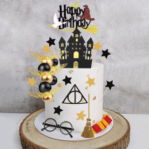 May include: A white cake decorated with a black castle, a Happy Birthday sign, a red witch hat, a white bird, gold wings, black and gold spheres, black stars, a triangle symbol, round black glasses, a small broom, and a red and yellow scarf.