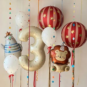 May include: A collection of party balloons including a large gold number three, two red and pink striped spheres with gold stars, a bear-shaped balloon, a lion-shaped balloon, and three white balloons with colorful confetti dots, all suspended with ribbons.