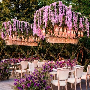 May include: Two rectangular crystal chandeliers suspended above a dining table, adorned with cascading purple wisteria flowers. The setup includes white upholstered chairs arranged around a table decorated with purple floral arrangements.