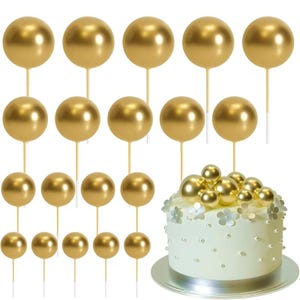 May include: Gold metallic sphere cake toppers on wooden sticks. The image shows a white frosted cake decorated with gold spheres and white flower accents. Additional gold sphere toppers are arranged around the cake.