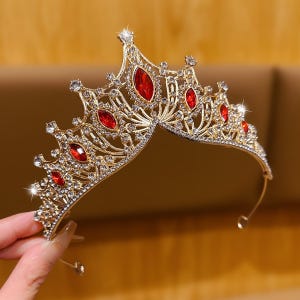 Peut inclure: Un diadème en métal doré présentant un design pointu et arqué, orné de nombreux strass transparents étincelants et de plusieurs cristaux rouges proéminents en forme de marquise, disposés symétriquement sur la structure en filigrane.