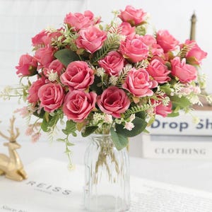 May include: A bouquet of artificial pink roses arranged in a clear glass vase. The arrangement features multiple rose blossoms in varying shades of pink, interspersed with green leaves and small white filler flowers, creating a dense floral display.