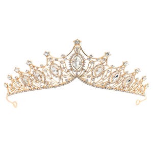 Peut inclure: Un diadème en métal doré présentant un design en filigrane complexe orné de nombreux cristaux transparents à facettes. La pièce comporte une pierre centrale ovale flanquée de cristaux plus petits en forme de marquise et ronds disposés symétriquement.