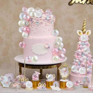 May include: A two-tiered pink cake decorated with iridescent bubble-shaped ornaments and floral accents. A smaller unicorn-themed cake is also present, along with various decorative elements like cookies and figurines, all in a pastel color scheme.