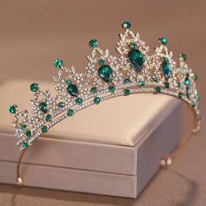 Peut inclure: Un diadème en métal doré présentant un motif complexe en filigrane rappelant des feuilles. La couronne est ornée de nombreuses petites pierres transparentes et de plusieurs pierres vert émeraude en forme de goutte d'eau.
