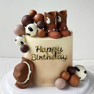 May include: A cylindrical cake with a light beige ombre finish, featuring a gold acrylic Happy Birthday topper. Decorations include brown cowboy boots, a brown cowboy hat, and clusters of brown and white cow-print spheres on top and base.
