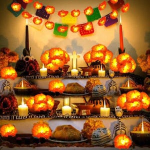 May include: A multi-tiered altar display featuring bright orange marigold flowers, lit pillar candles, and small skeleton figurines. A string of colourful paper banners hangs above, while round loaves of bread are placed on plates across the levels.
