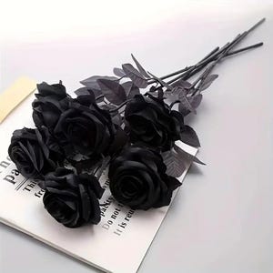 May include: A bouquet of six black artificial roses with dark green stems and leaves. The roses are in full bloom and arranged on a white surface with text from a book visible.