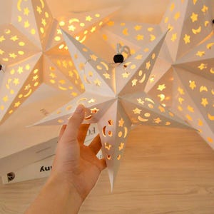 May include: A white star-shaped paper lantern featuring intricate cutouts of stars and crescent moons. The lantern is illuminated from within, casting a warm glow through the patterns. A small black bead is attached to the top hanging cord.