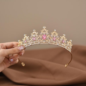 Peut inclure: Un diadème doré présentant un design symétrique avec un filigrane métallique complexe, des strass transparents étincelants et cinq cristaux roses en forme de goutte disposés le long du bord supérieur arqué.