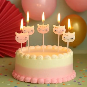 May include: A layered birthday cake with pink and cream frosting, adorned with five lit cat-shaped candles. The candles feature closed eyes and whiskers. The background includes pink and gold balloons and confetti.