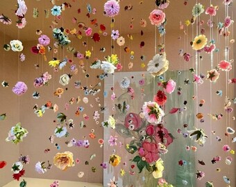May include: A room decorated with numerous colorful flowers suspended from the ceiling. The flowers, in shades of pink, yellow, white, and purple, create a whimsical and artistic display. A painting of flowers is visible in the background.