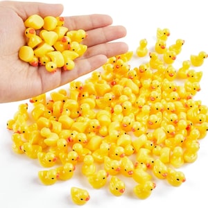 May include: A collection of small, yellow duck figurines with orange beaks and eyes. Some are held in a hand, while others are scattered on a white surface. The ducks are made of a glossy material and are a cheerful yellow color.
