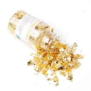 Peut inclure: Un pot en verre transparent de feuilles d'or, dont une partie du contenu est renversée sur une surface blanche. Le pot est étiqueté "Decorativ Gold Foil". Les feuilles d'or sont en flocons irréguliers et chatoyants.
