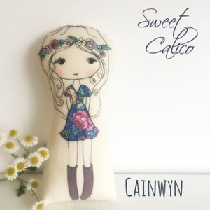Op de afbeelding: Een handgemaakte pop met een bloemenjurk en een bloemenkroon. De pop heeft bruine ogen en een lieve uitdrukking. De pop is gemaakt van stof en heeft een witte achtergrond. De tekst "Sweet Calico" en "CAINWYN" is zichtbaar op de afbeelding.