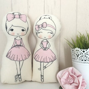 May include: Two handmade fabric dolls dressed as ballerinas. Both dolls have pink tutus, black hair, and black eyes. The doll on the left has a pink bow in her hair, while the doll on the right has a pink headband.