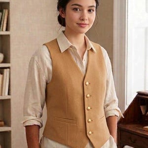 May include: A tan vest with gold buttons over a white collared shirt. The vest has two front pockets and a tailored fit. The woman is wearing the vest and shirt in a room with a bookshelf and a desk.